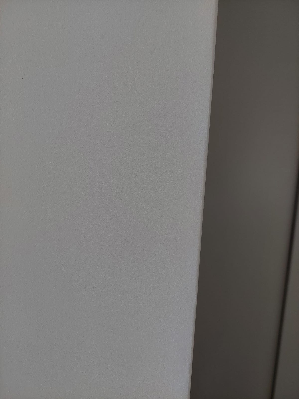 Fragment ściany pomalowanej na jasnoszaro, z widoczną fakturą i niewielką ciemną plamką, sąsiadującej z ciemniejszą, niewyraźną powierzchnią.
