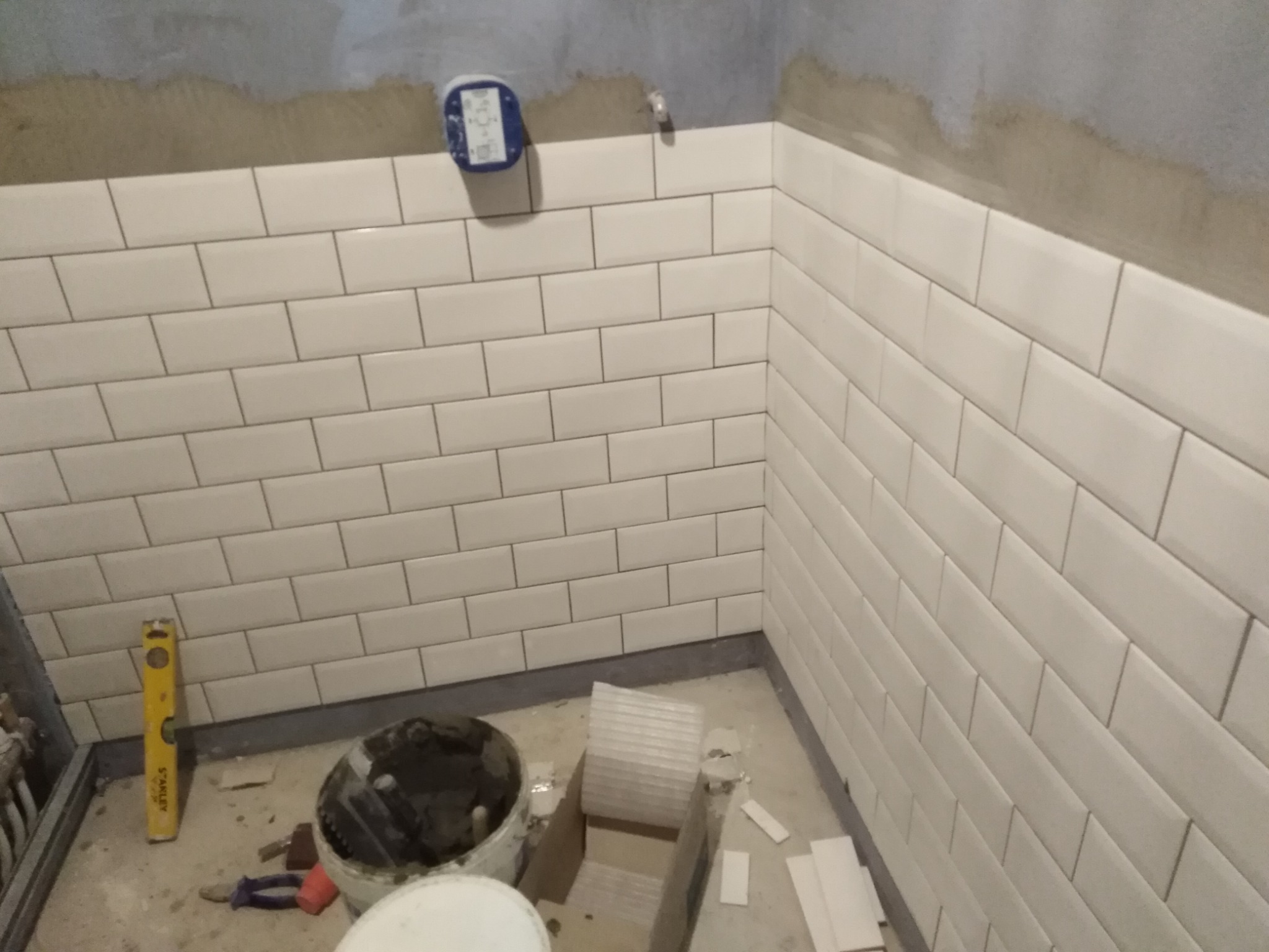 Ściana łazienki w trakcie układania białych, prostokątnych płytek typu metro, z widocznym poziomem, wiaderkiem z zaprawą i fragmentem niezakończonej ściany powyżej płytek.