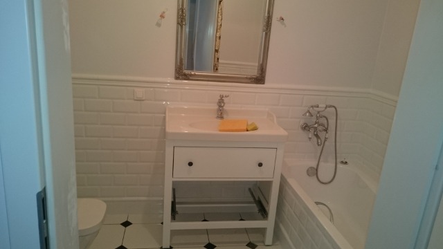 Biała łazienka po odświeżeniu z umywalką na szafce z otwartą szufladą, baterią, mydłem, lustrem w ozdobnej ramie i częściowo widoczną wanną z baterią.