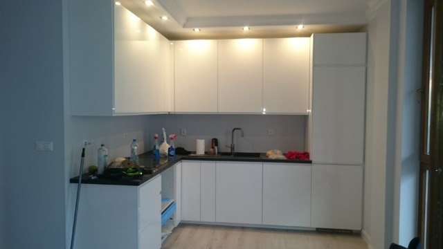 Nowoczesna kuchnia z białymi, błyszczącymi szafkami podświetlonymi punktowymi lampami sufitowymi, czarny blat i widoczne środki czystości sugerują świeżo zakończony montaż.