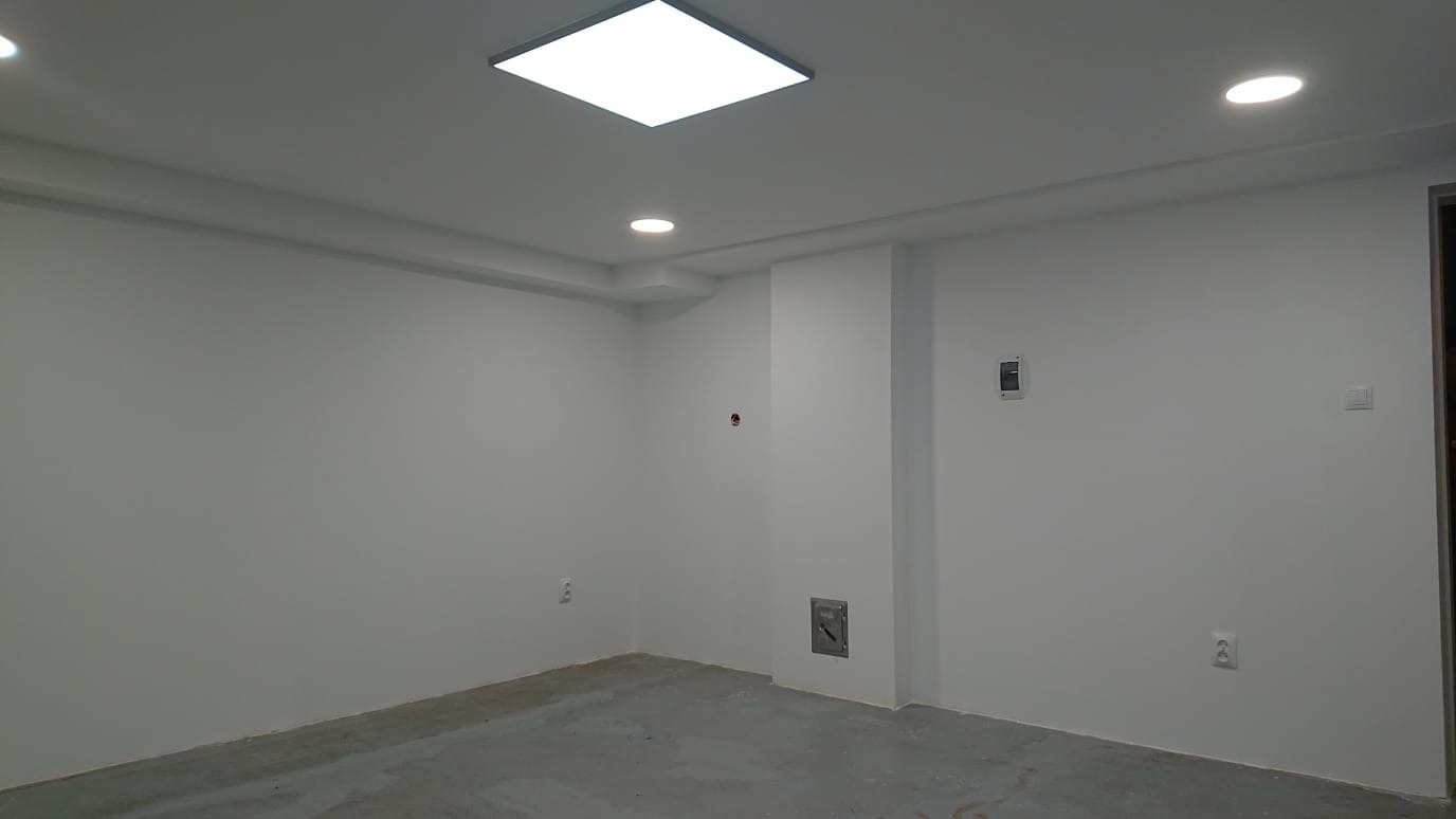 Pomalowane na biało puste pomieszczenie z betonową podłogą, oświetleniem sufitowym i gniazdkami elektrycznymi na ścianach.