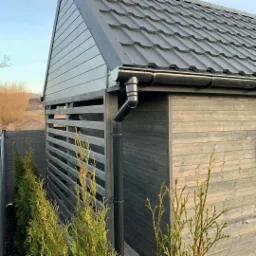 Zadaszona konstrukcja z ciemnoszarej blachy dachowej i drewnianych ścian z poziomymi listwami, z czarną rynną i rurą spustową, otoczona tujami.