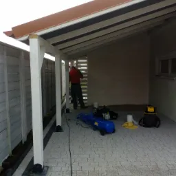 Konstrukcja zadaszenia na tarasie w trakcie montażu, widoczny pracownik w czerwonej bluzie, narzędzia i materiały budowlane na kostce brukowej.