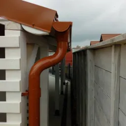 Brązowa rynna spustowa zamontowana przy białej, ażurowej ścianie budynku, widok z dołu na tle pochmurnego nieba.