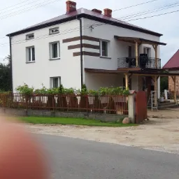 Dwupiętrowy dom z białą elewacją i brązowymi pasami dekoracyjnymi, balkon z czarną balustradą, widoczna drabina i wiadro, ogrodzenie z czerwonej cegły, niebo z chmurami.