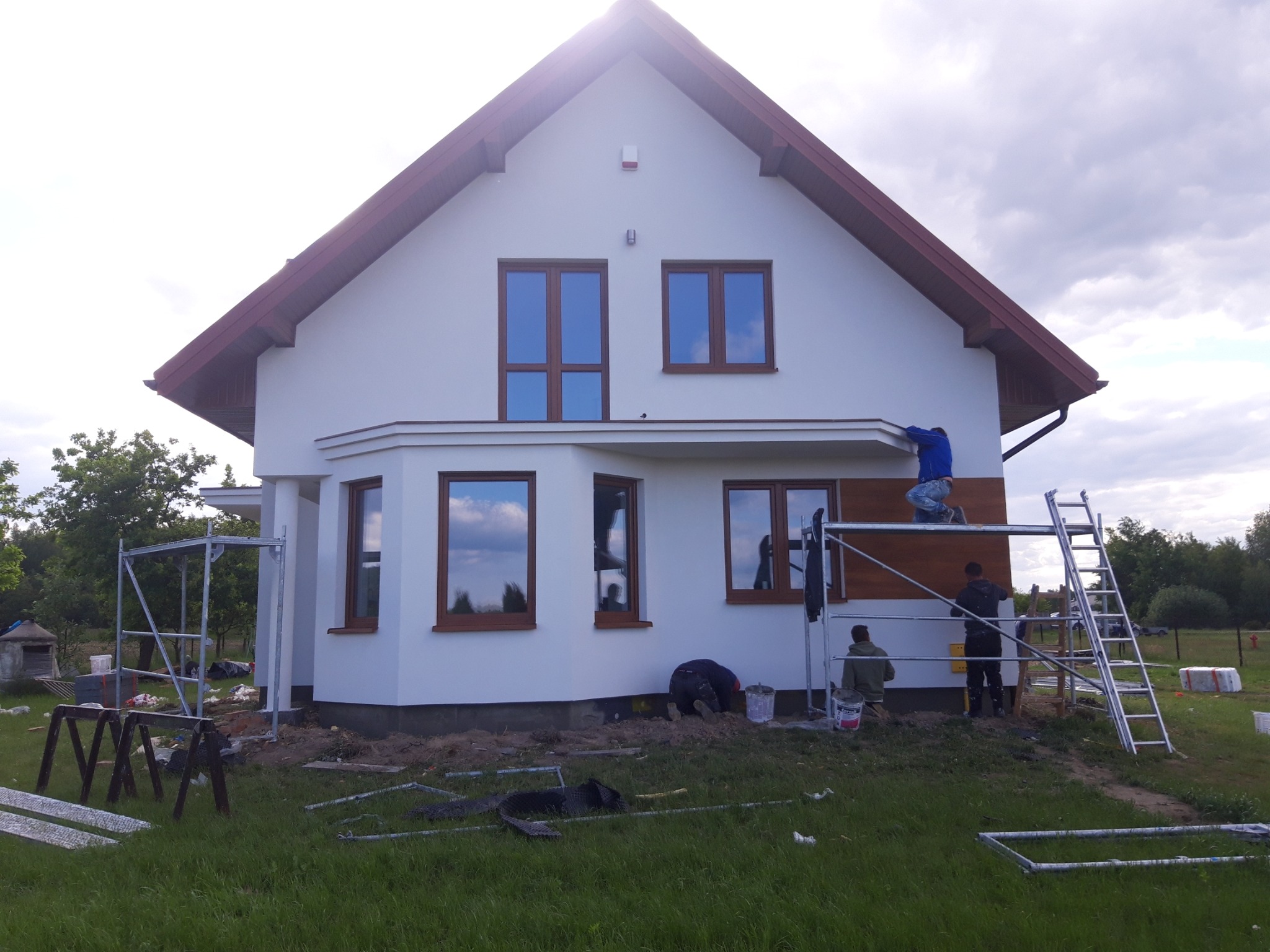 Montaż drewnianej elewacji na białym domu jednorodzinnym z brązowymi oknami, widoczne rusztowania i pracownicy w trakcie pracy.