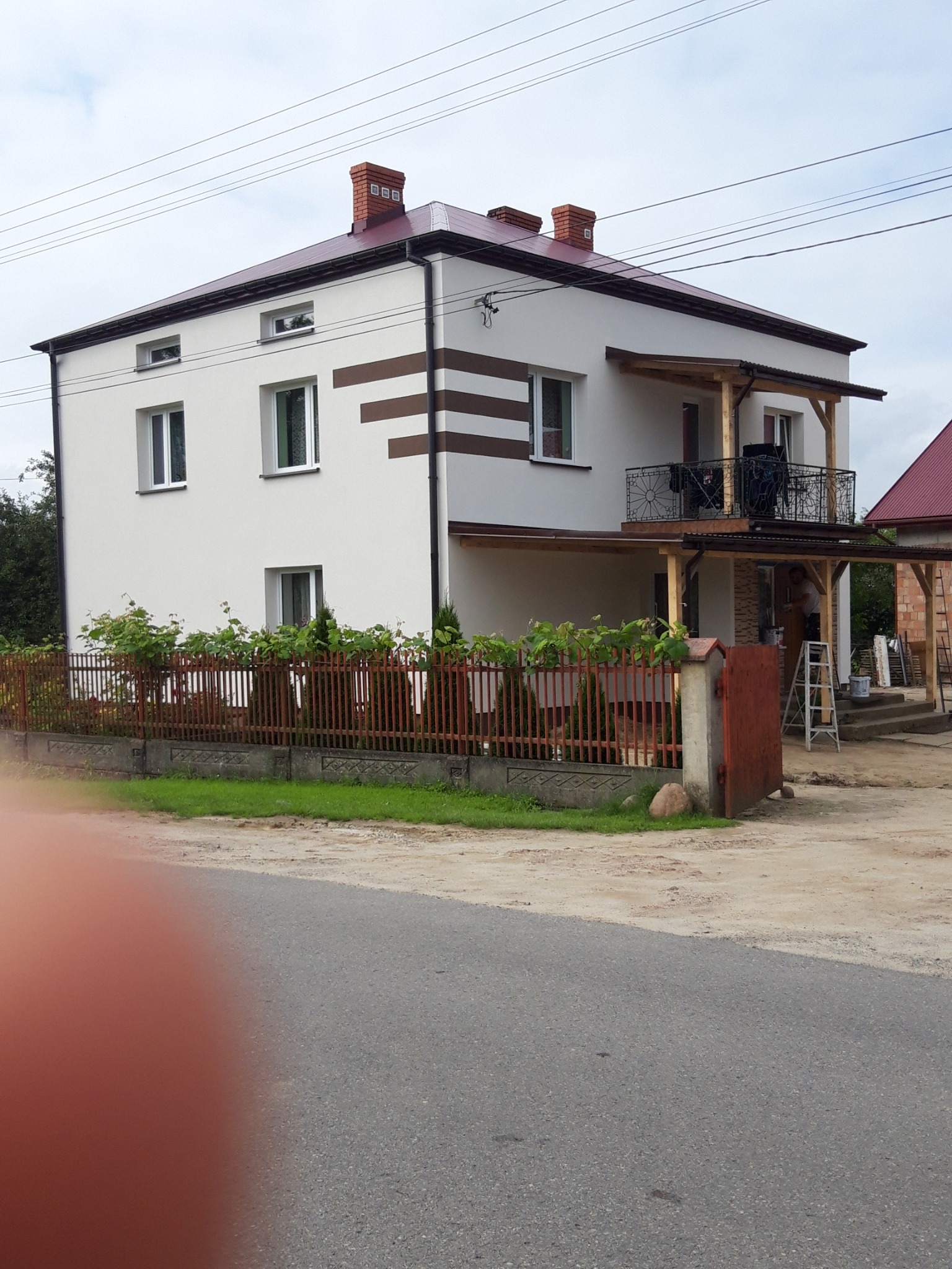 Dwupiętrowy dom z białą elewacją i brązowymi pasami dekoracyjnymi, balkon z czarną balustradą, widoczna drabina i wiadro, ogrodzenie z czerwonej cegły, niebo z chmurami.