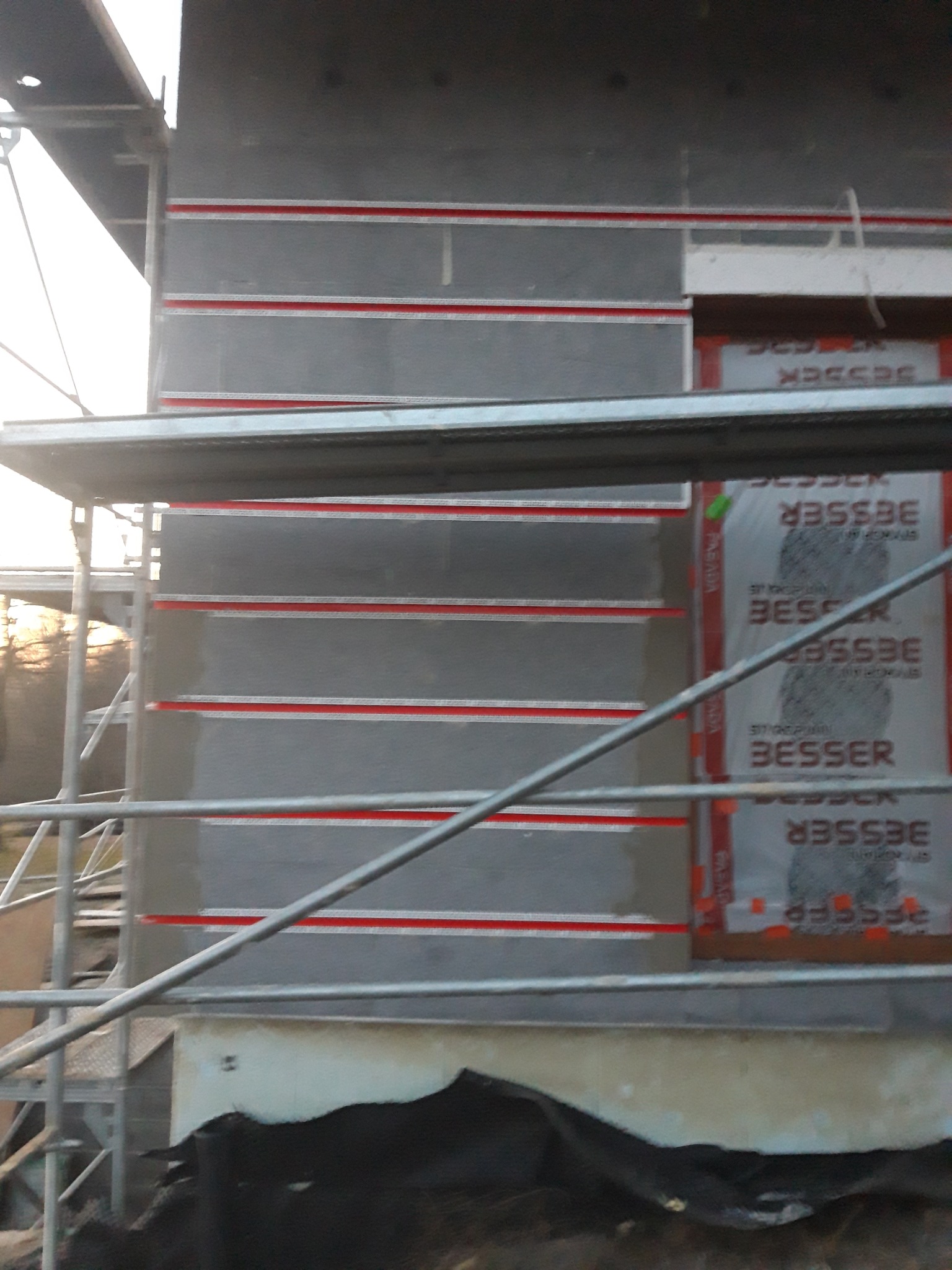 Fragment elewacji budynku w trakcie prac, widoczne rusztowania, płyty izolacyjne z widocznym logo producenta i poziomymi czerwonymi paskami, częściowo pokryte warstwą szarego materiału.