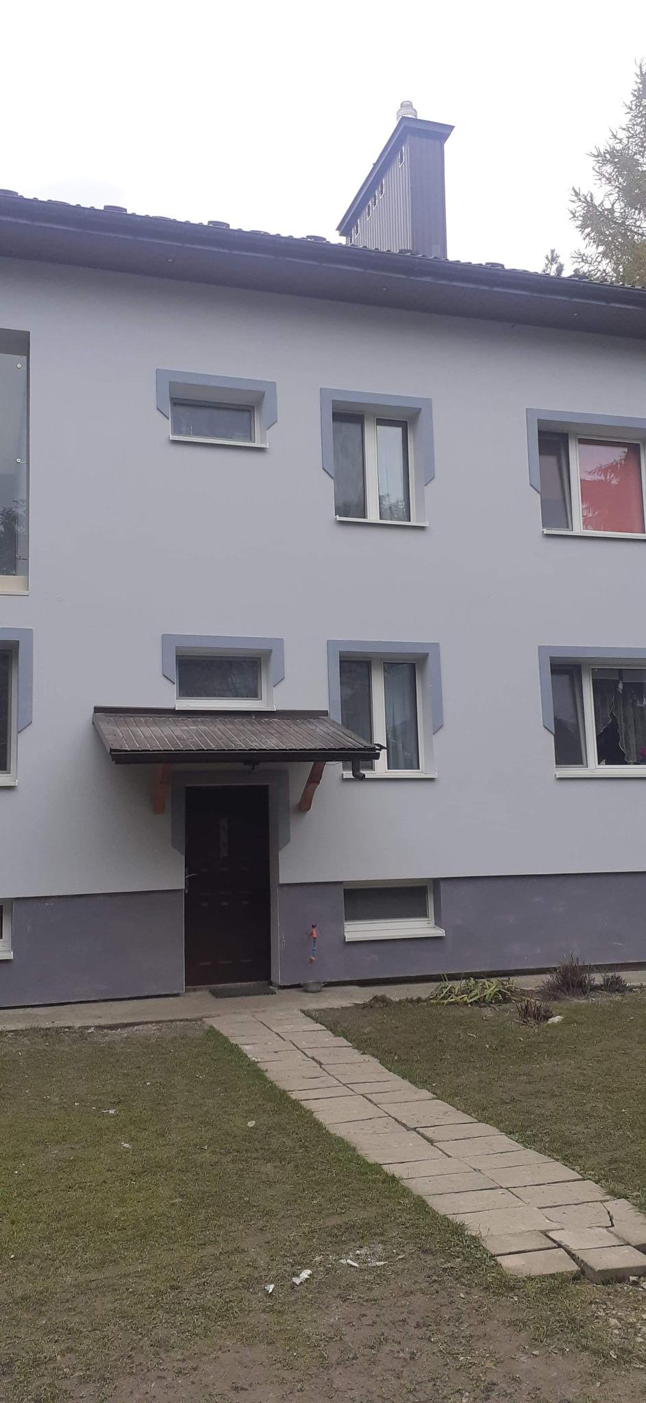 Dwupiętrowy budynek mieszkalny z szarą elewacją, ciemną podmurówką i charakterystycznymi obramowaniami okien w odcieniu szarości, widziany z poziomu ścieżki prowadzącej do wejścia.