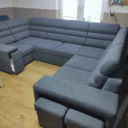 Szary, modułowy narożnik z funkcją spania i pojemnikiem na pościel, widoczne dwa pufy, na podłodze panele, w tle okno i warsztat.