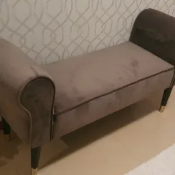 Stylowa, tapicerowana ławka w odcieniach szarości z ciemnymi nóżkami zakończonymi złotymi akcentami, ustawiona w pokoju z wzorzystą tapetą i rośliną doniczkową.