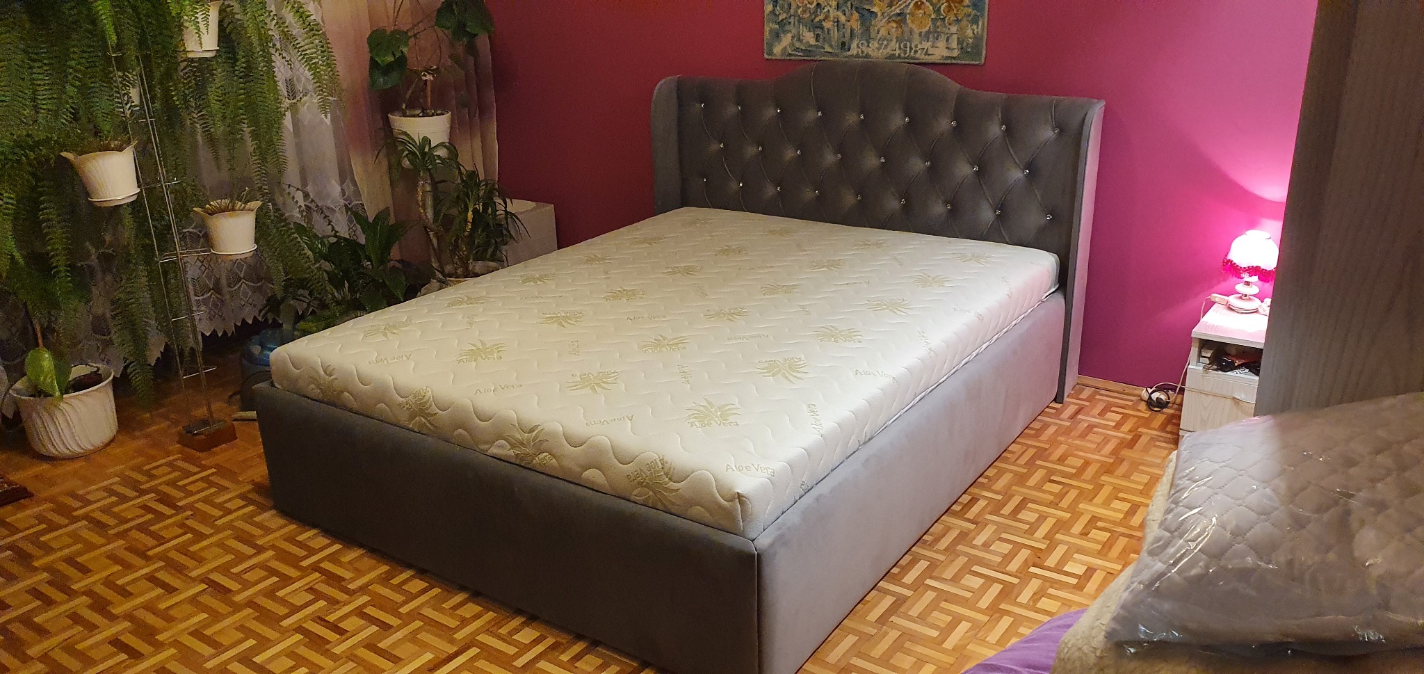 Szare tapicerowane łóżko z pikowanym zagłówkiem i materacem Aloe Vera w sypialni z parkietem i roślinami doniczkowymi.