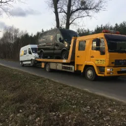 Żółta autolaweta MAN przewozi na platformie uszkodzonego, ciemnego busa UPS; w tle biały bus GLS na drodze asfaltowej.