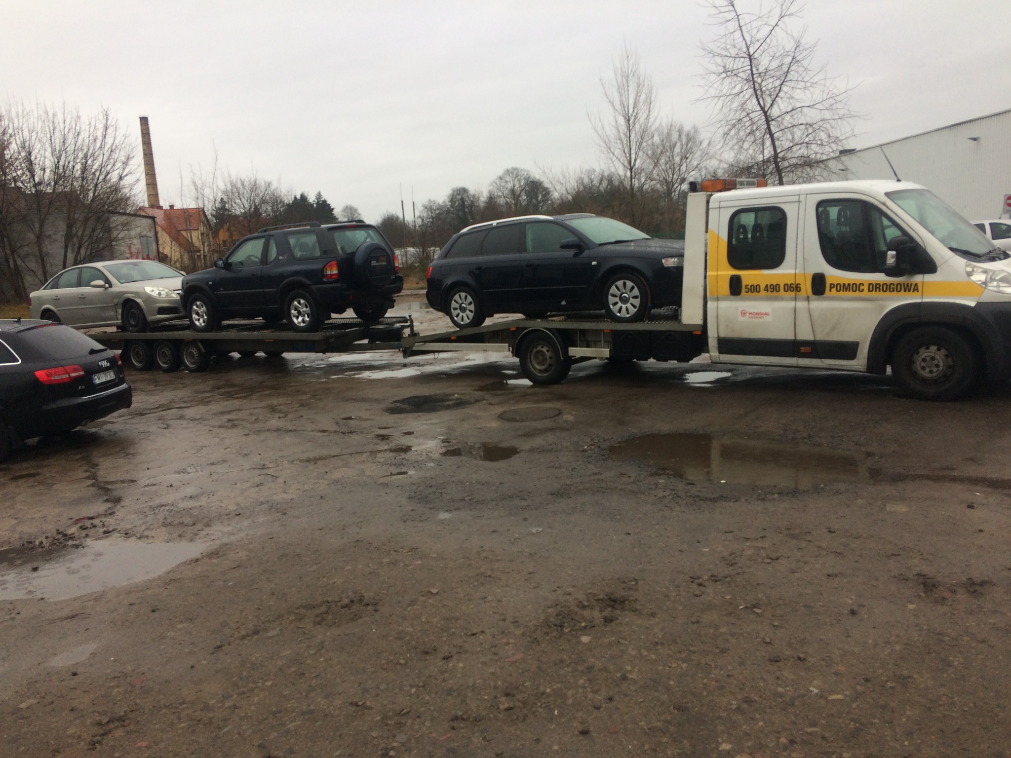 Laweta z trzema samochodami osobowymi na przyczepie, zaparkowana na błotnistym placu, widoczna reklama 'POMOC DROGOWA' na pojeździe