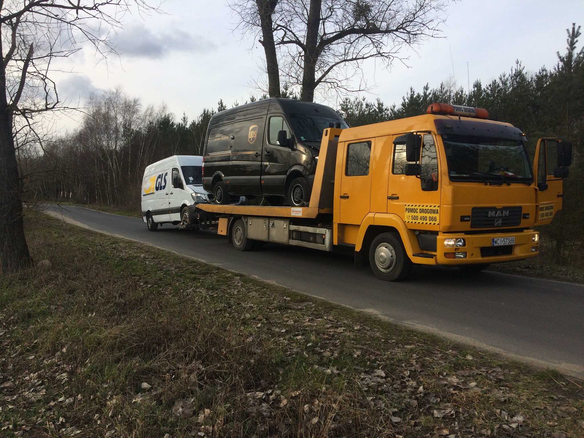 Żółta autolaweta MAN przewozi na platformie uszkodzonego, ciemnego busa UPS; w tle biały bus GLS na drodze asfaltowej.