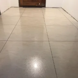 Ułożona podłoga z jasnych, kwadratowych płytek z ciemnymi fugami, widoczne fragmenty ścian i drewniane drzwi w tle, ślady po pracach wykończeniowych przy ścianach.