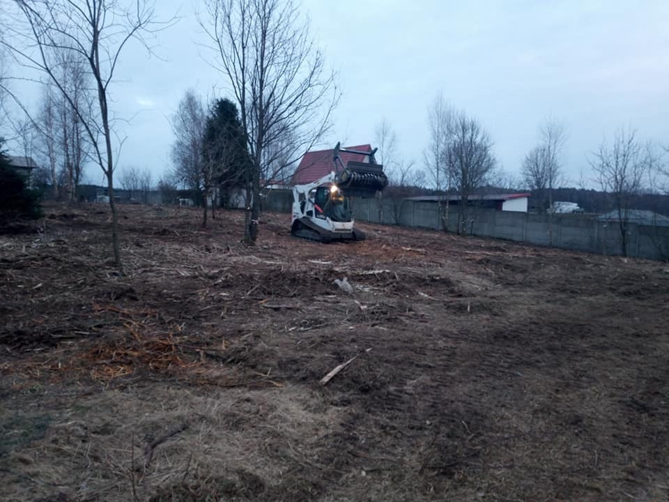 Bobcat z chwytakiem usuwa pozostałości po wycince drzew na rozległym terenie, widoczny fragment domu z czerwoną dachówką i betonowe ogrodzenie w tle.