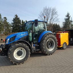 POWER ENERGI SEBASTIAN JUSZCZAK - Niebieski traktor New Holland z żółtą przyczepą na kostce brukowej, w tle drzewa i błękitne niebo. Widok z boku, opony z wyraźnym bieżnikiem.