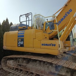 POWER ENERGI SEBASTIAN JUSZCZAK - Żółta koparka Komatsu PC210LC z bliska, z widocznymi gąsienicami i elementami konstrukcyjnymi, na tle zielonych drzew i błękitnego nieba.