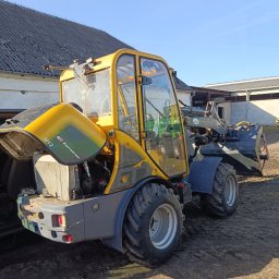 POWER ENERGI SEBASTIAN JUSZCZAK - Żółto-szara ładowarka teleskopowa Eurotrac W12F z otwartą maską silnika, stojąca na ziemi przed budynkiem gospodarczym w słoneczny dzień. Widoczne detale konstrukcyjne.