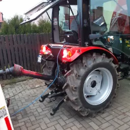 Czerwony ciągnik z otwartą kabiną i podłączonym wałem odbioru mocy, widoczne tylne światła i tablica rejestracyjna, na pierwszym planie kostka brukowa.