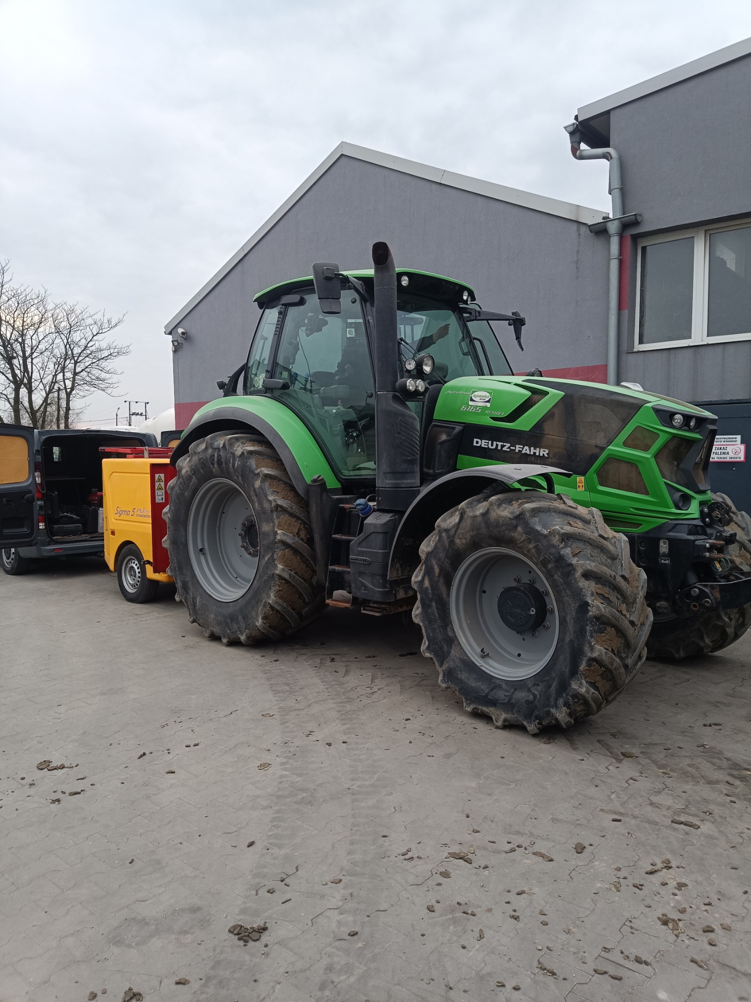 Zielony traktor Deutz-Fahr z przyczepką i otwartym busem w tle, stojący na betonowym placu przed szarym budynkiem. Widoczne błoto na oponach.