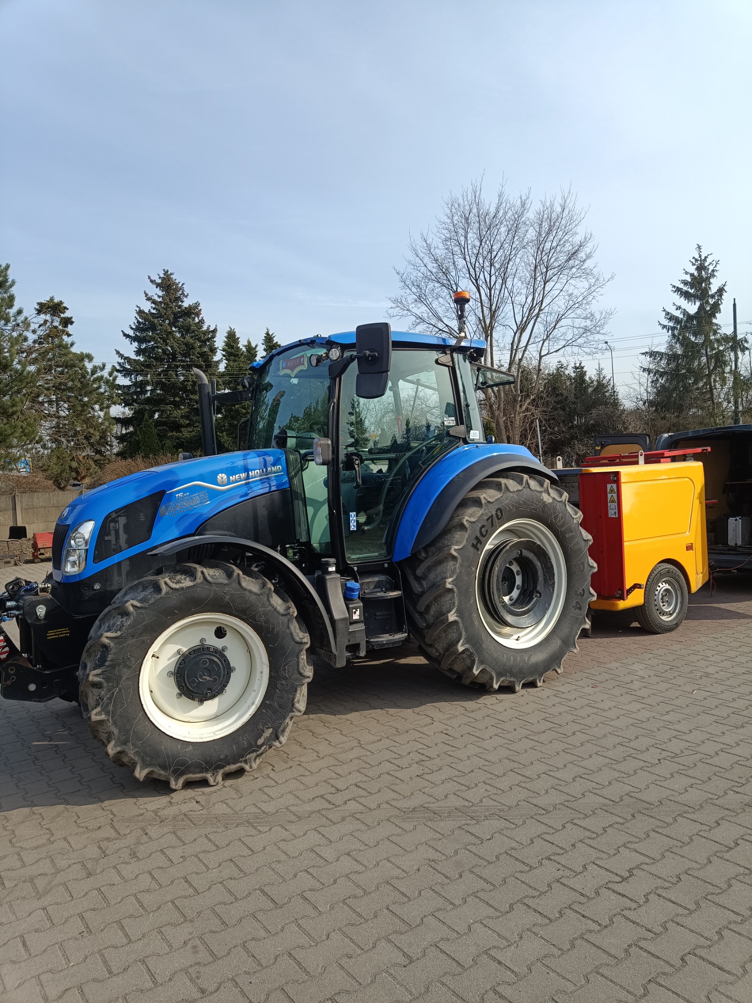Niebieski traktor New Holland z żółtą przyczepą na kostce brukowej, w tle drzewa i błękitne niebo. Widok z boku, opony z wyraźnym bieżnikiem.