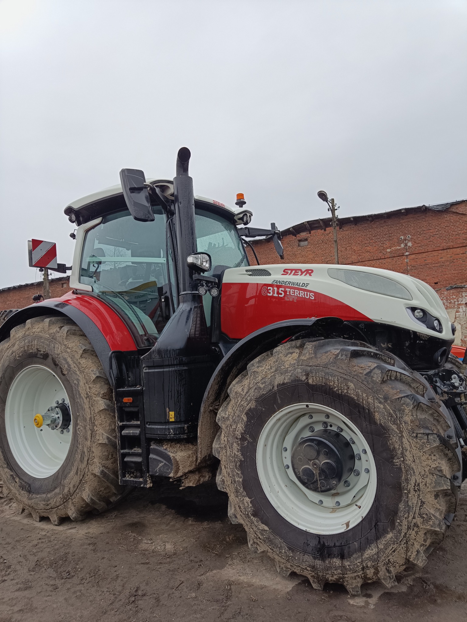 Czerwono-biały traktor Steyr 6315 Terrus z dużymi, ubłoconymi kołami na tle ceglanego budynku i pochmurnego nieba. Widok z dołu, podkreślający masywność maszyny.