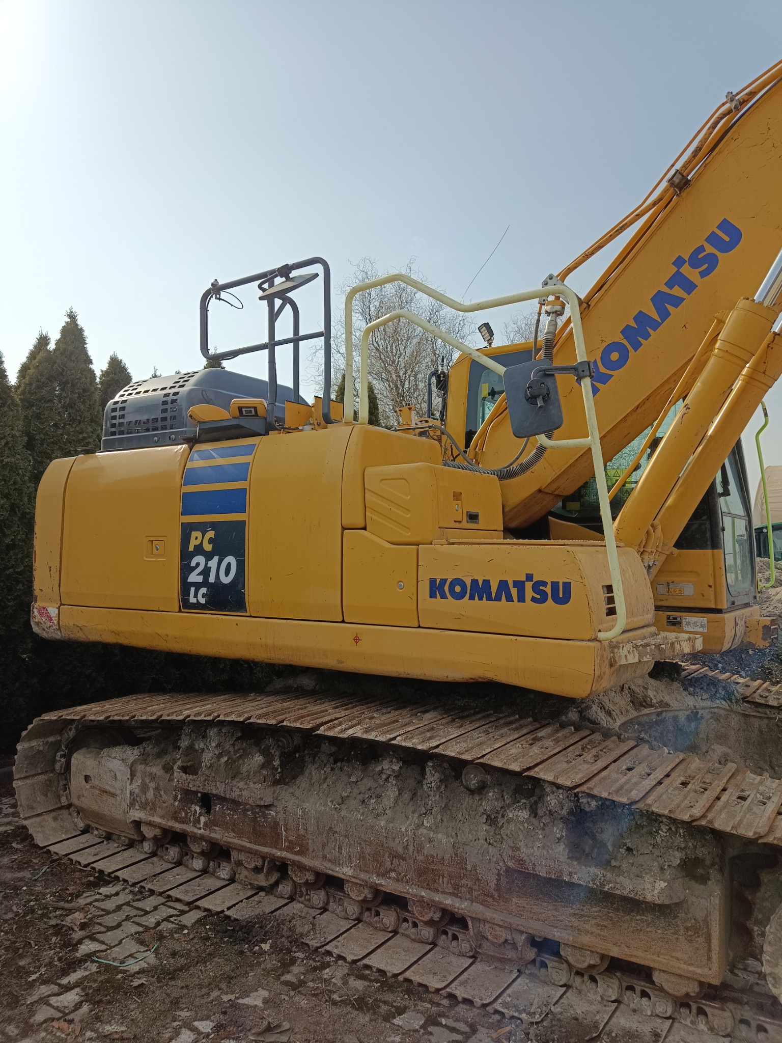 Żółta koparka Komatsu PC210LC z bliska, z widocznymi gąsienicami i elementami konstrukcyjnymi, na tle zielonych drzew i błękitnego nieba.