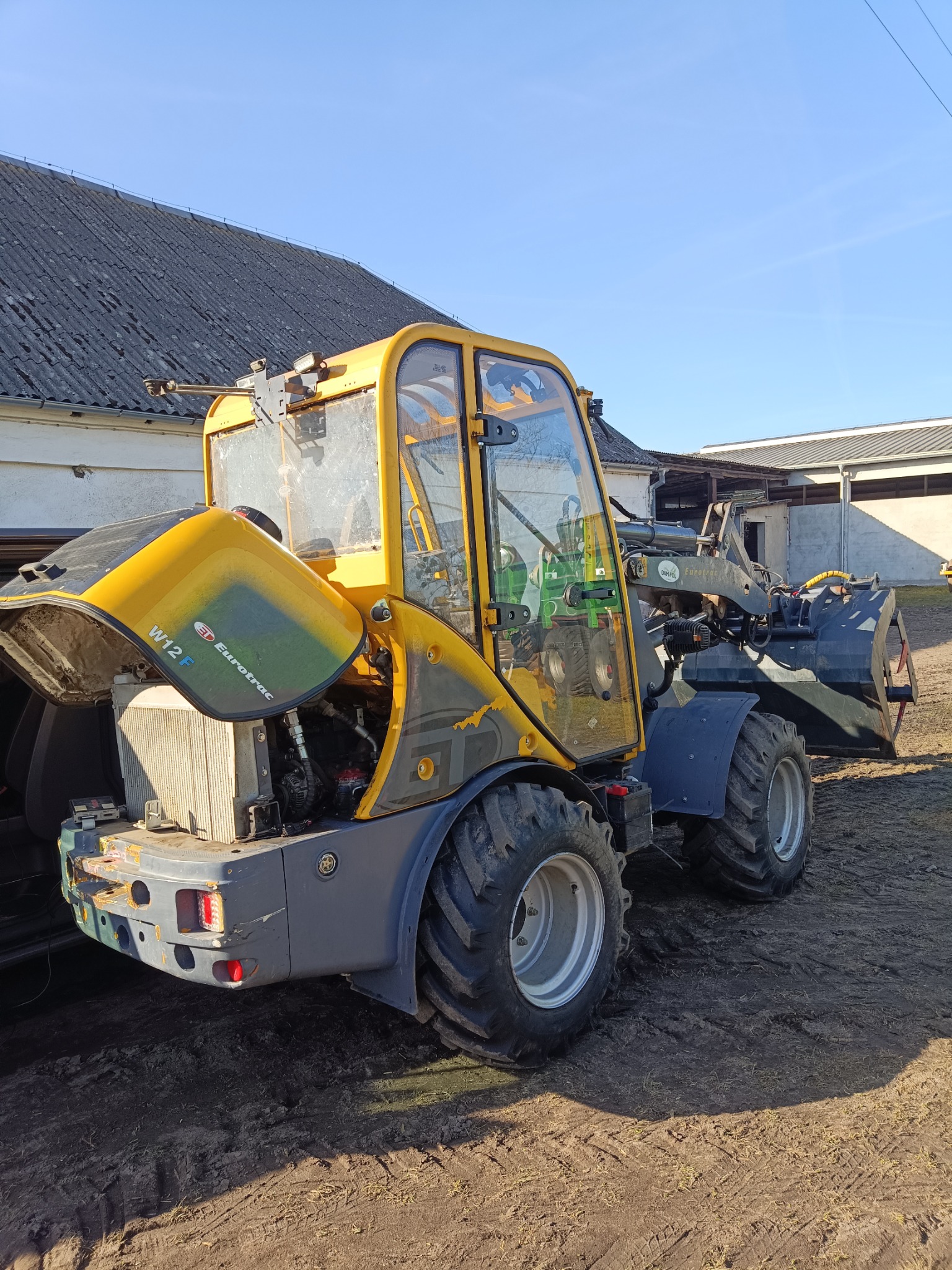 Żółto-szara ładowarka teleskopowa Eurotrac W12F z otwartą maską silnika, stojąca na ziemi przed budynkiem gospodarczym w słoneczny dzień. Widoczne detale konstrukcyjne.