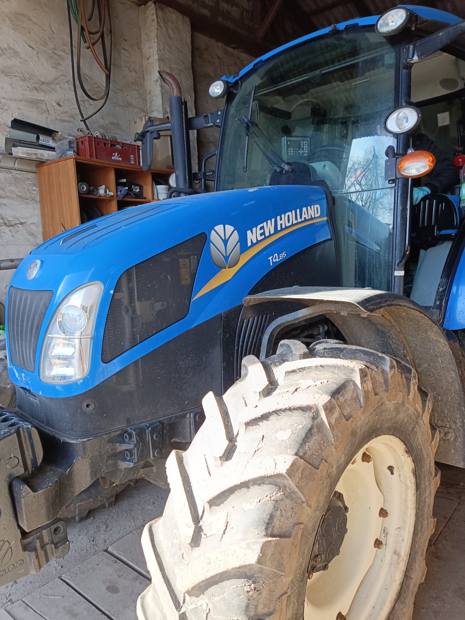 Niebieski traktor New Holland T4.85 w garażu, widok z przodu na oponę i maskę. Widoczne detale: reflektory, logo i oznaczenia modelu. W tle półki z narzędziami.