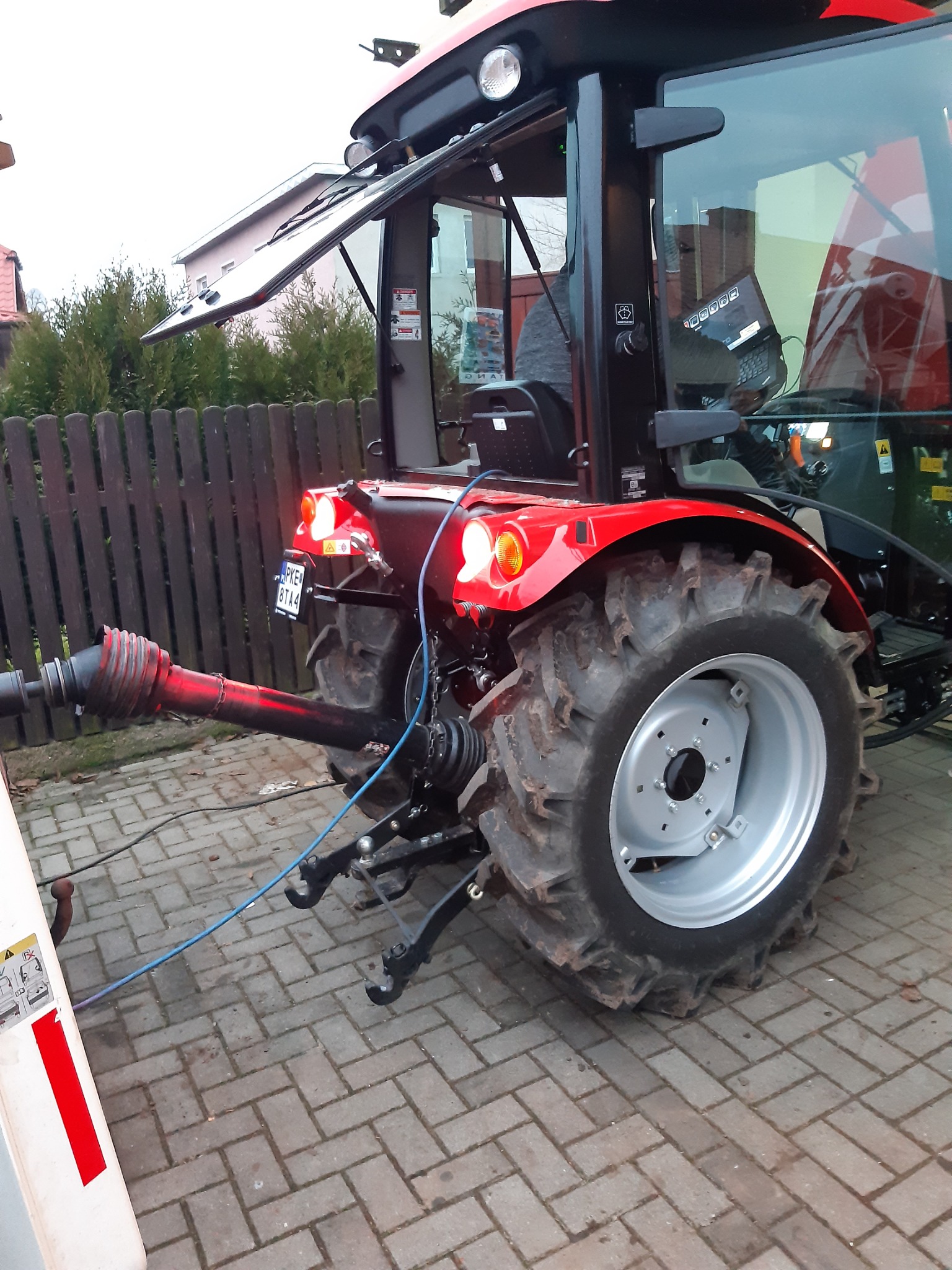 Czerwony ciągnik z otwartą kabiną i podłączonym wałem odbioru mocy, widoczne tylne światła i tablica rejestracyjna, na pierwszym planie kostka brukowa.