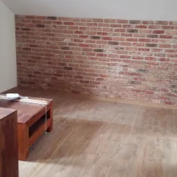 Wykończona ściana z cegły dekoracyjnej w poddaszowym pokoju w Krakowie, z drewnianą podłogą i stolikiem kawowym w rogu.