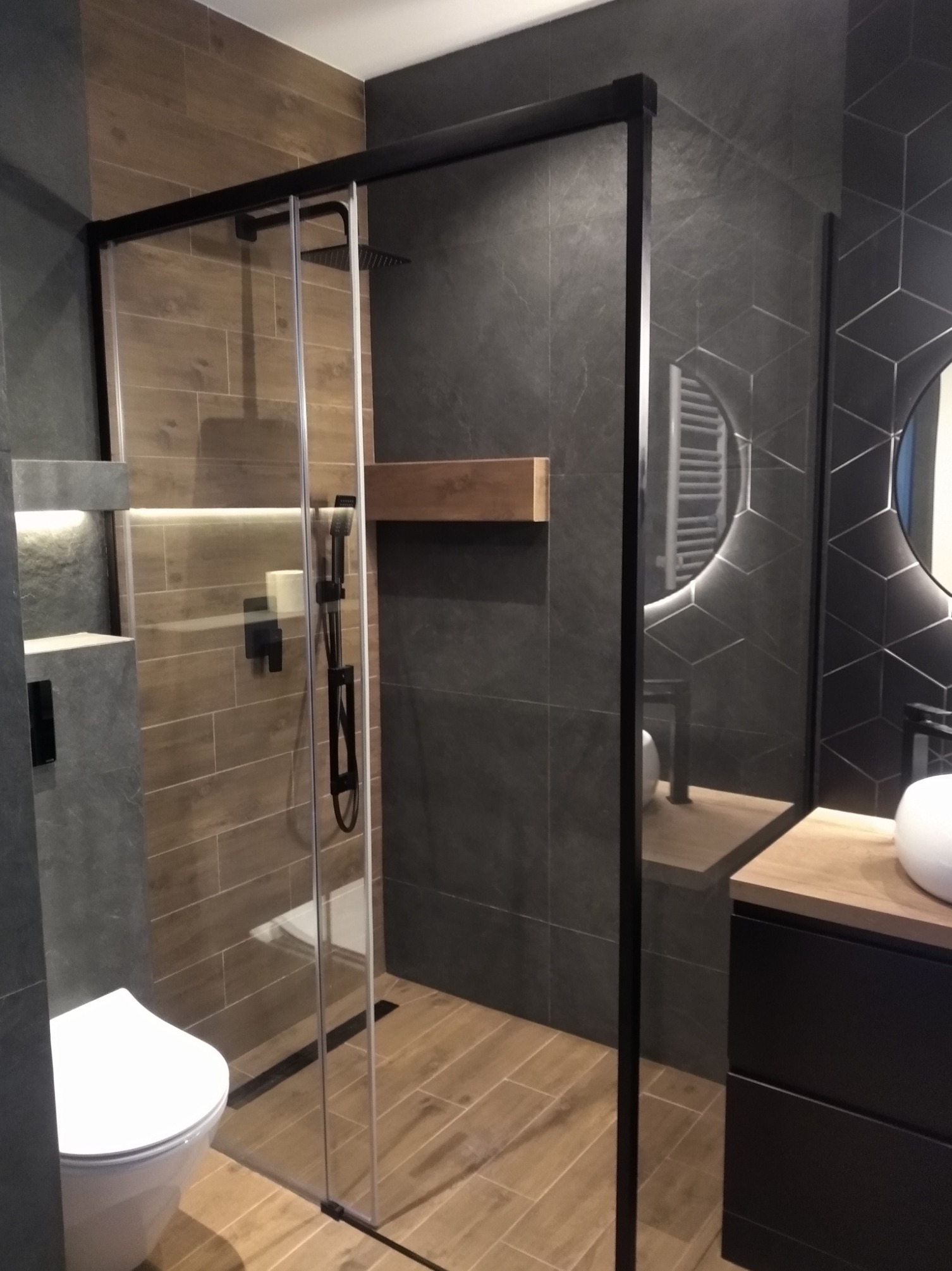 Nowoczesna łazienka z kabiną prysznicową typu walk-in z czarnymi profilami, drewnopodobnymi płytkami i geometryczną mozaiką na ścianie.