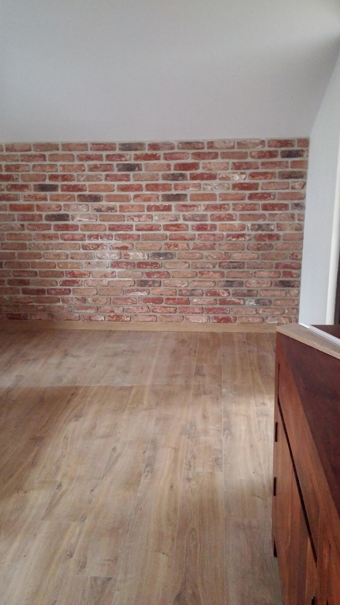 Ściana z cegły klinkierowej w odcieniach czerwieni i brązu, na poddaszu ze skośnym sufitem, połączona z drewnianą podłogą i fragmentem mebla z ciemnego drewna.