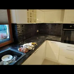 Czarna, trójwymiarowa mozaika ułożona na ścianie nad blatem w kuchni, białe szafki, widoczny zlew z naczyniami, silikon i pistolet do silikonu na blacie.