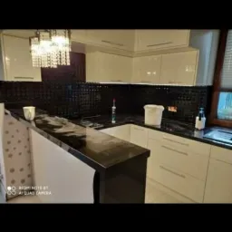 Wykończona kuchnia z białymi szafkami, czarnym blatem i mozaiką nad blatem, widoczny fragment ściany z tapetą w kropki i wiszący żyrandol.