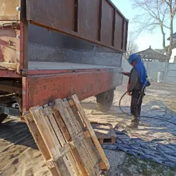 Przygotowanie do malowania przyczepy rolniczej: Pracownik w kombinezonie ochronnym piaskuje rdzawą powierzchnię, z boku leżą drewniane palety.