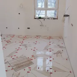 Układanie płytek podłogowych imitujących marmur w jasnej łazience, z użyciem czerwonych krzyżyków dystansowych, widok na okno z zimowym krajobrazem.