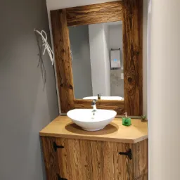 Rustykalna łazienka z drewnianą szafką pod umywalkę, lustro w ramie z postarzanego drewna i białą umywalką nablatową.