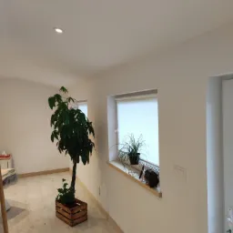 Jasne wnętrze po odświeżeniu: białe ściany, drewniana podłoga, roślina w drewnianej skrzynce i doniczka na parapecie okna z roletą.