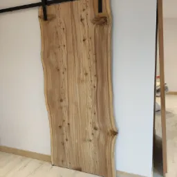 Drewniane drzwi przesuwne z naturalną krawędzią, zawieszone na czarnym, metalowym systemie szynowym, widoczne na tle białej ściany i jasnej podłogi.