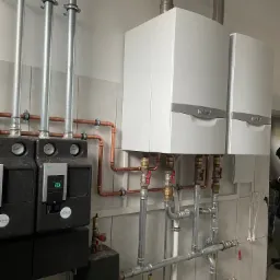 Dwa białe piece grzewcze Vaillant zamontowane na ścianie z szarymi płytkami, widoczne rury miedziane i stalowe, oraz czarne pompy obiegowe Wilo i Concept.