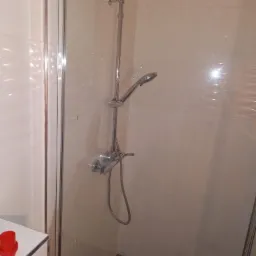 Nowoczesna kabina prysznicowa z chromowaną armaturą i beżowymi płytkami, widoczny fragment półki z czerwonym pojemnikiem.