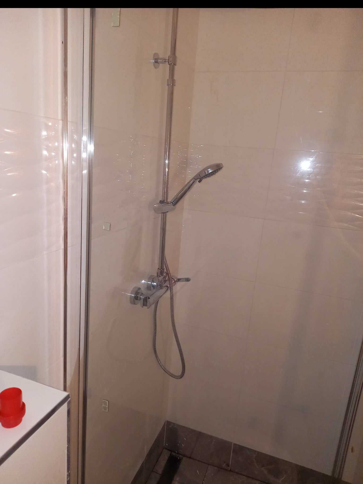 Nowoczesna kabina prysznicowa z chromowaną armaturą i beżowymi płytkami, widoczny fragment półki z czerwonym pojemnikiem.