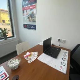 Biuro agenta ubezpieczeniowego: plakat Superpolisa z ofertą ubezpieczeń domu i mieszkania na ścianie, biurko z laptopem, kalendarzem firmowym, ulotkami i miseczką cukierków.