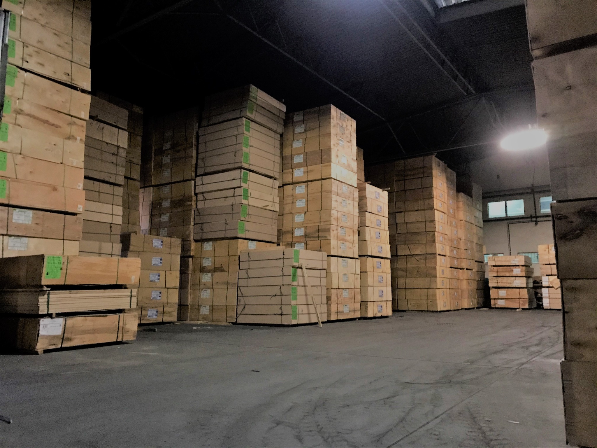 Magazyn z wysokimi stertami zapakowanych płyt drewnianych i sklejki, oświetlony pojedynczą lampą sufitową.