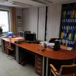 Biuro z drewnianym biurkiem, dwoma komputerami, stosem dokumentów, regałem pełnym segregatorów i dwoma krzesłami na kółkach.