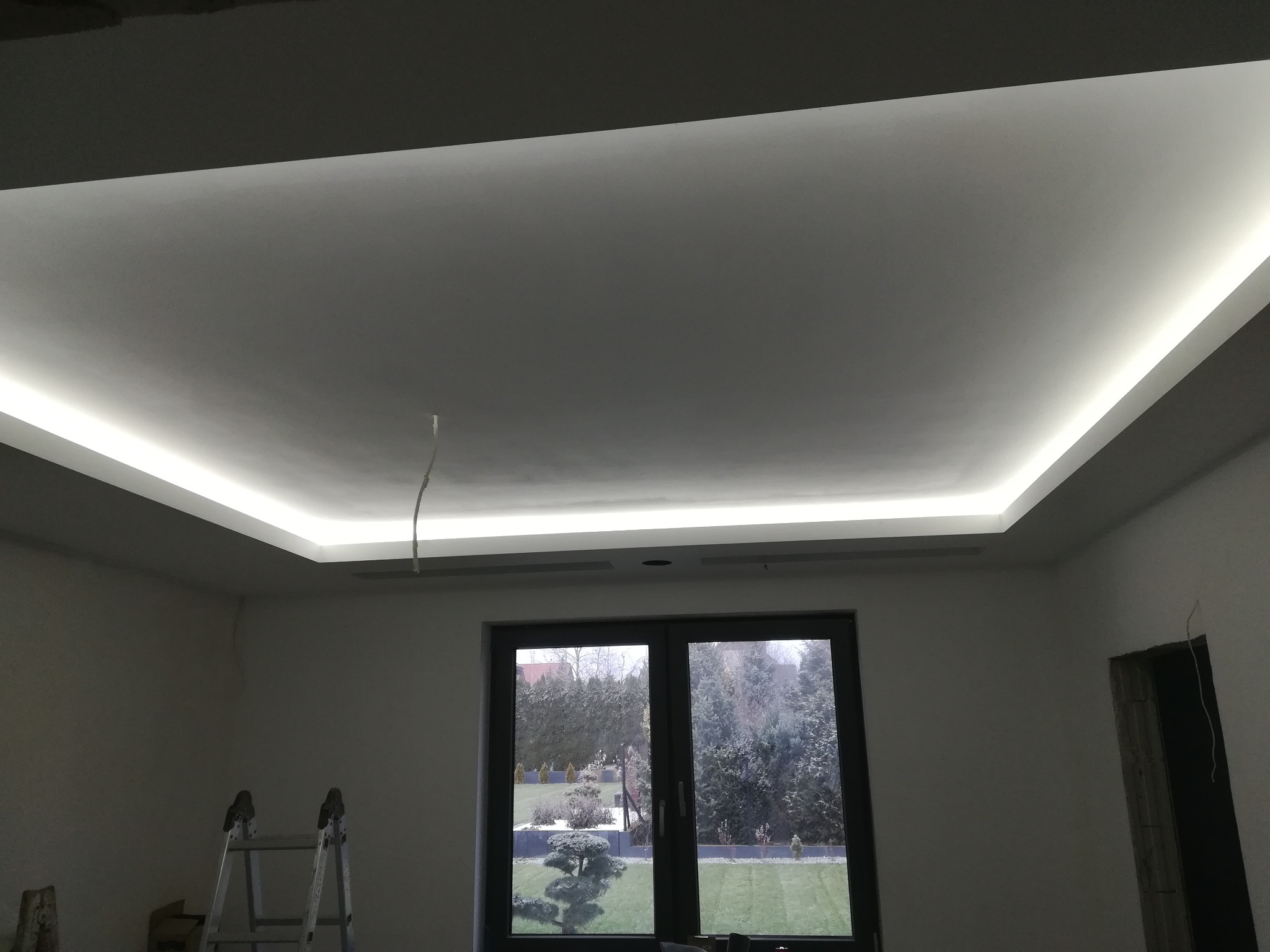 Podwieszany sufit z oświetleniem LED w nowo wykończonym pokoju, widoczne drabiny malarskie i okno z widokiem na ogród.