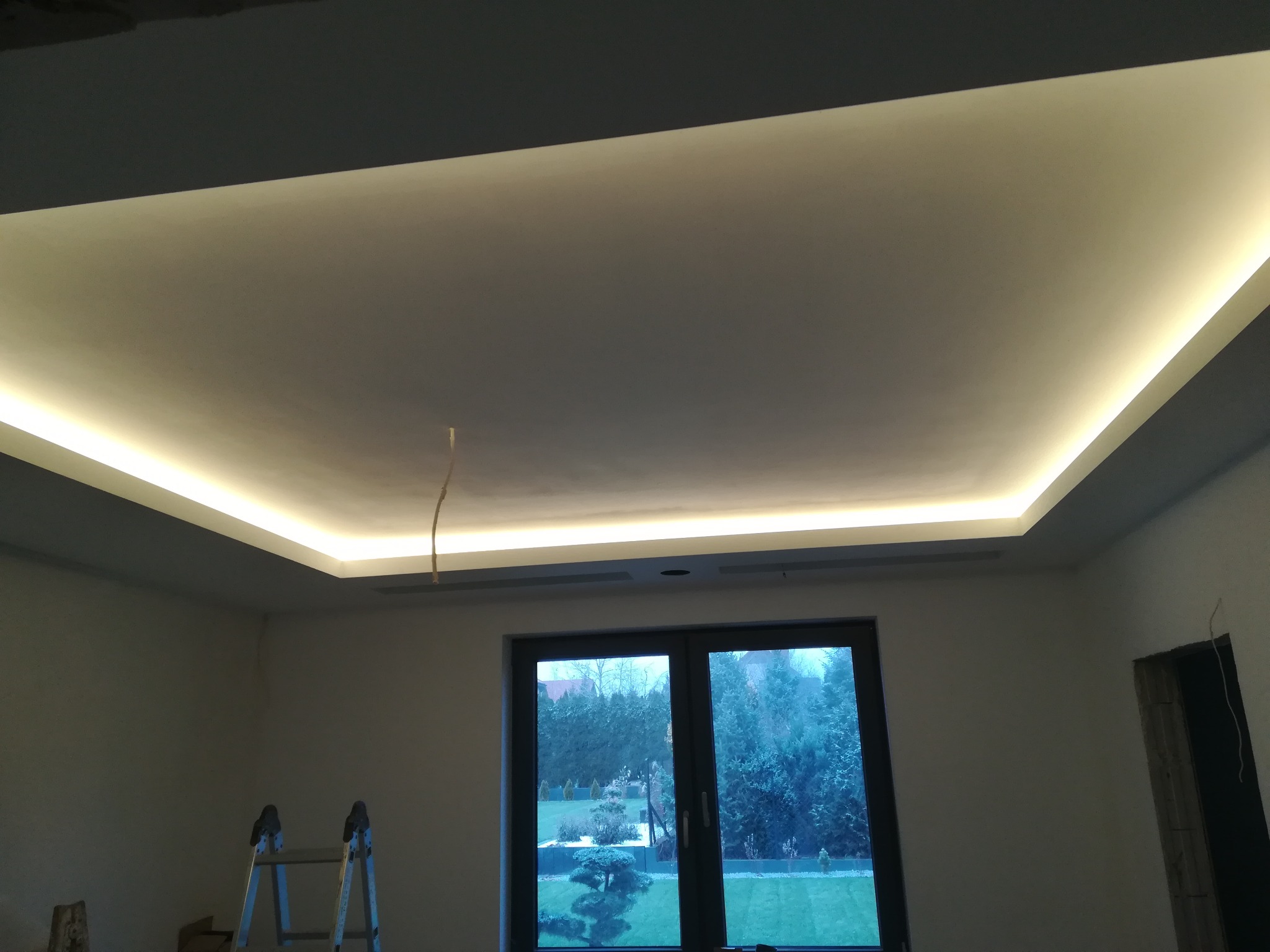 Nowoczesny sufit podwieszany z oświetleniem LED, widoczna drabina malarska i okno z widokiem na ogród